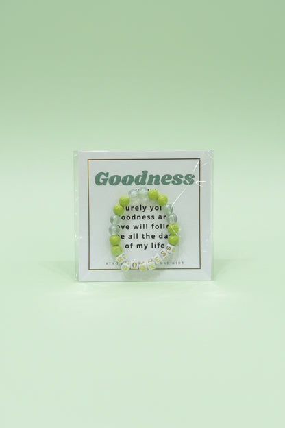 A hand holds a packaged beaded bracelet on a printed card that reads 'Goodness' in green text, with a pink and cream boutique display blurred in the background.