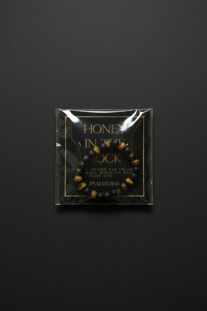 A hand holding a packaged beaded bracelet with a black background card that reads 'HONEY IN THE ROCK' and 'PSALMS 81:6'. The bracelet itself appears dark brown or black with small gold and red accents.