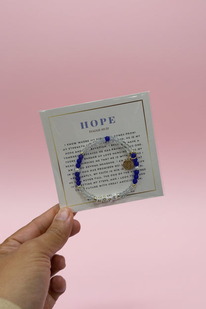 A hand holds a small jewelry display card featuring a delicate bracelet with blue beads and gold-toned charms, set against a softly blurred pink retail display background.