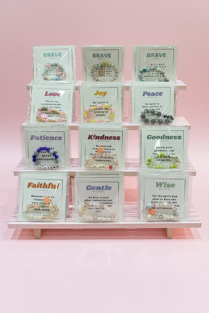A display of multiple small acrylic cards, each holding a beaded bracelet in a clear plastic sleeve. Each card has a different inspirational word or phrase printed on it, such as Brave, Love, Joy, Peace, Patience, Kindness, Goodness, Faithful, Gentle, and Wise.