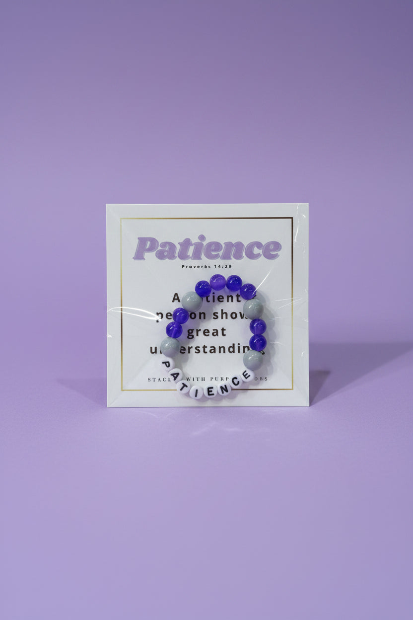 A hand holds a clear plastic card displaying a beaded bracelet with purple and gray beads and a silver letter charm spelling 'PATIENCE'. The background is a softly blurred pink jewelry display.