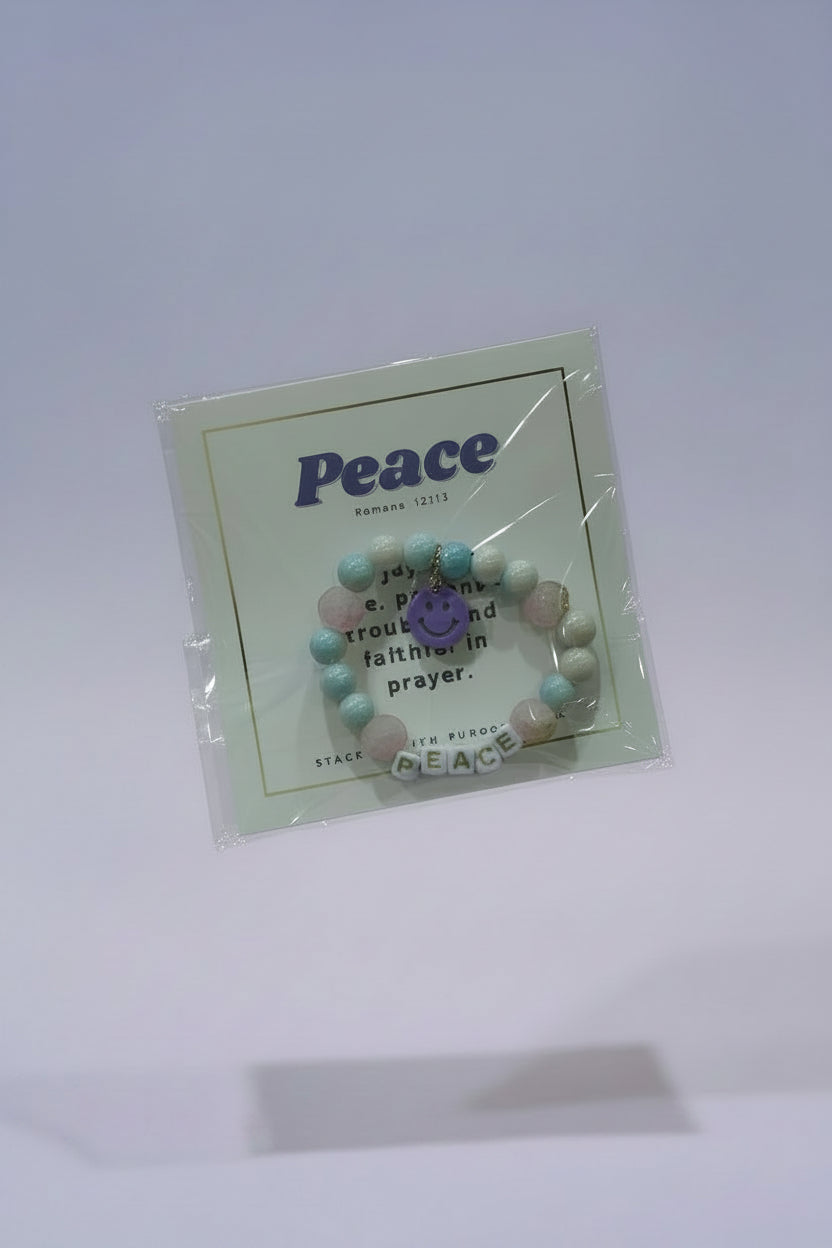 A hand holds a packaged beaded bracelet on a white backing card printed with the word "Peace" and a small purple peace sign. The bracelet contains pastel pink, light blue, and white beads with a purple center charm.