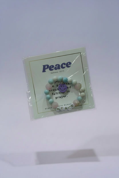 A hand holds a packaged beaded bracelet on a white backing card printed with the word "Peace" and a small purple peace sign. The bracelet contains pastel pink, light blue, and white beads with a purple center charm.