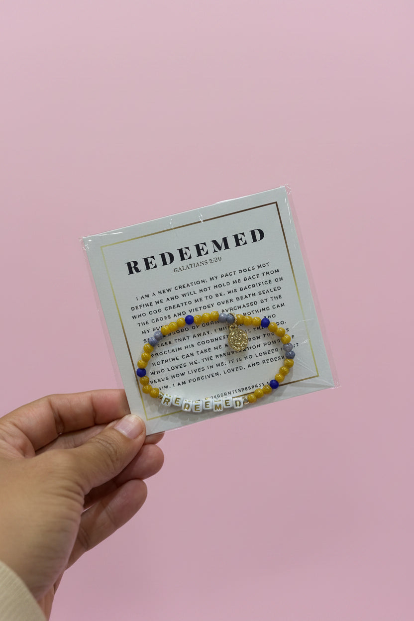 A hand holds a small jewelry display card featuring a beaded bracelet with a gold-toned charm. The card reads 'REDEEMED' and 'Calvin Klein', and the background shows a pink boutique display with other bracelets and boxes.