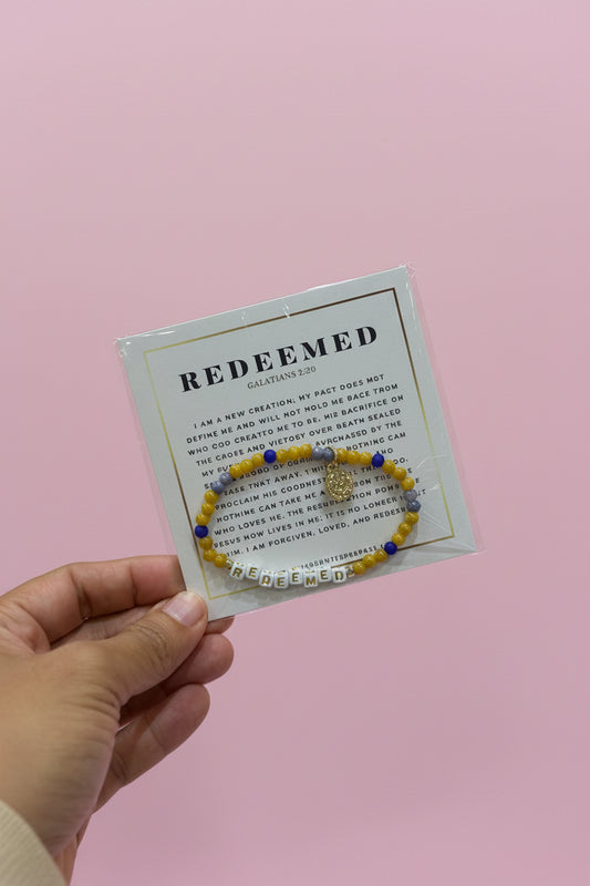 A hand holds a small jewelry display card featuring a beaded bracelet with a gold-toned charm. The card reads 'REDEEMED' and 'Calvin Klein', and the background shows a pink boutique display with other bracelets and boxes.