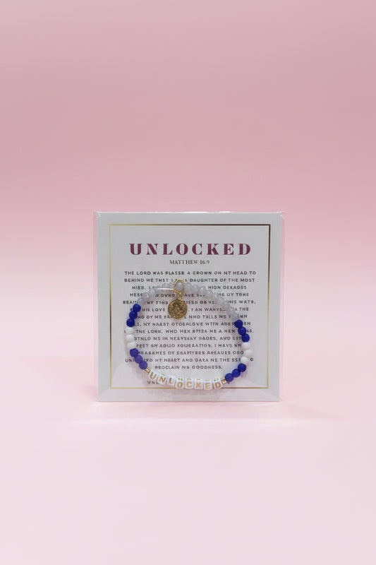 A hand holds a small jewelry display card featuring a beaded bracelet with a gold-toned heart charm. The card is branded 'UNLOCKED' and is photographed in a pink boutique setting with blurred jewelry displays in the background.